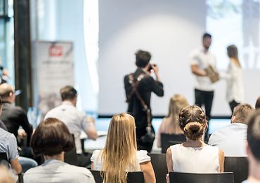 Publikum hört einem Vortrag in einem Konferenzraum mit Präsentation auf Leinwand zu.